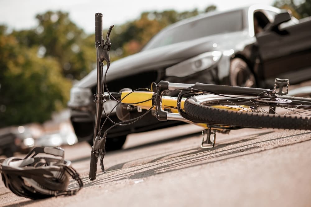 A yellow and black bicycle lies on the ground next to a dark SUV with an open door, suggesting a recent accident, with a helmet visible nearby.