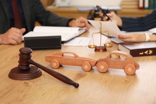 Wooden toy cars representing a collision on a lawyer’s desk with a gavel, scales of justice, and legal documents in the background.